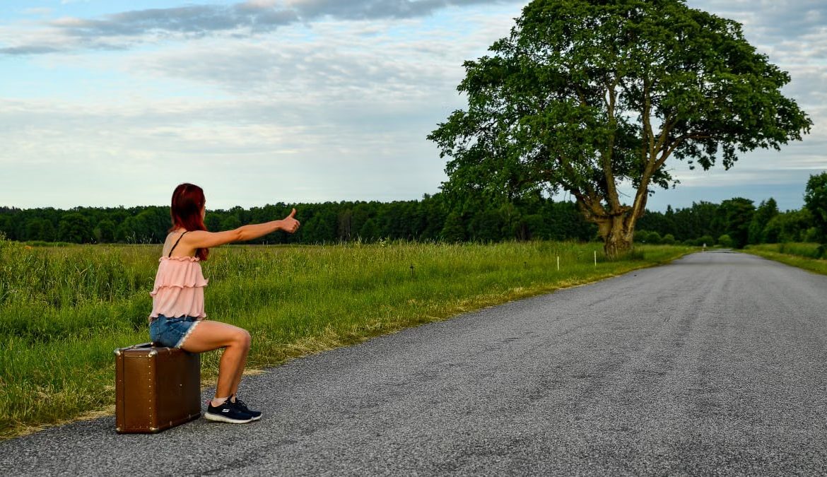 A woman sits on a suitcase, hitchhiking along a peaceful country road in Kolga, Estonia.